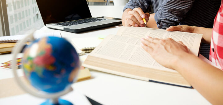 Two people study at a bright desk with an open book, a laptop, a pencil, and a globe in the foreground.