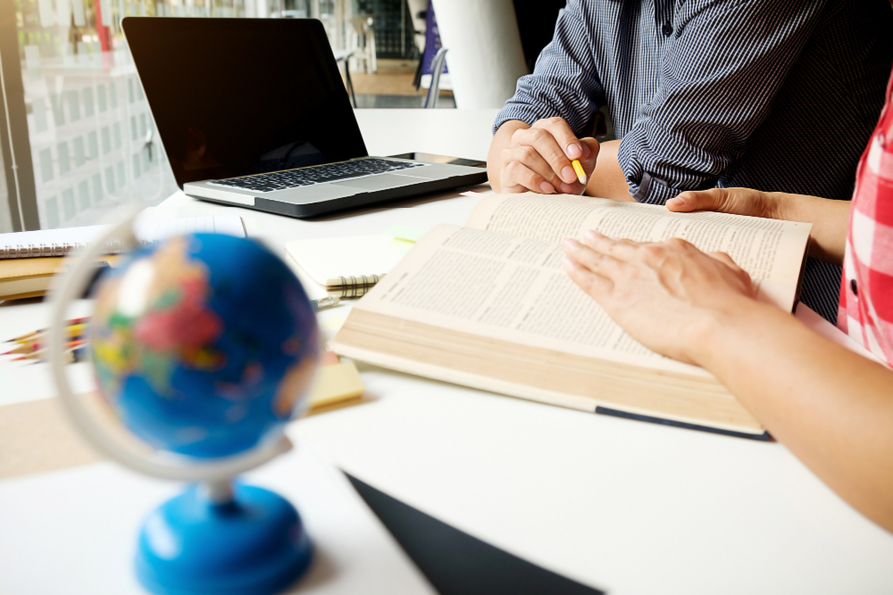 Two people study at a bright desk with an open book, a laptop, a pencil, and a globe in the foreground.