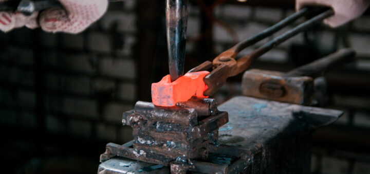 Hot orange metal being shaped in a blacksmith's hammering vise, with gloved hands and tongs in a workshop.