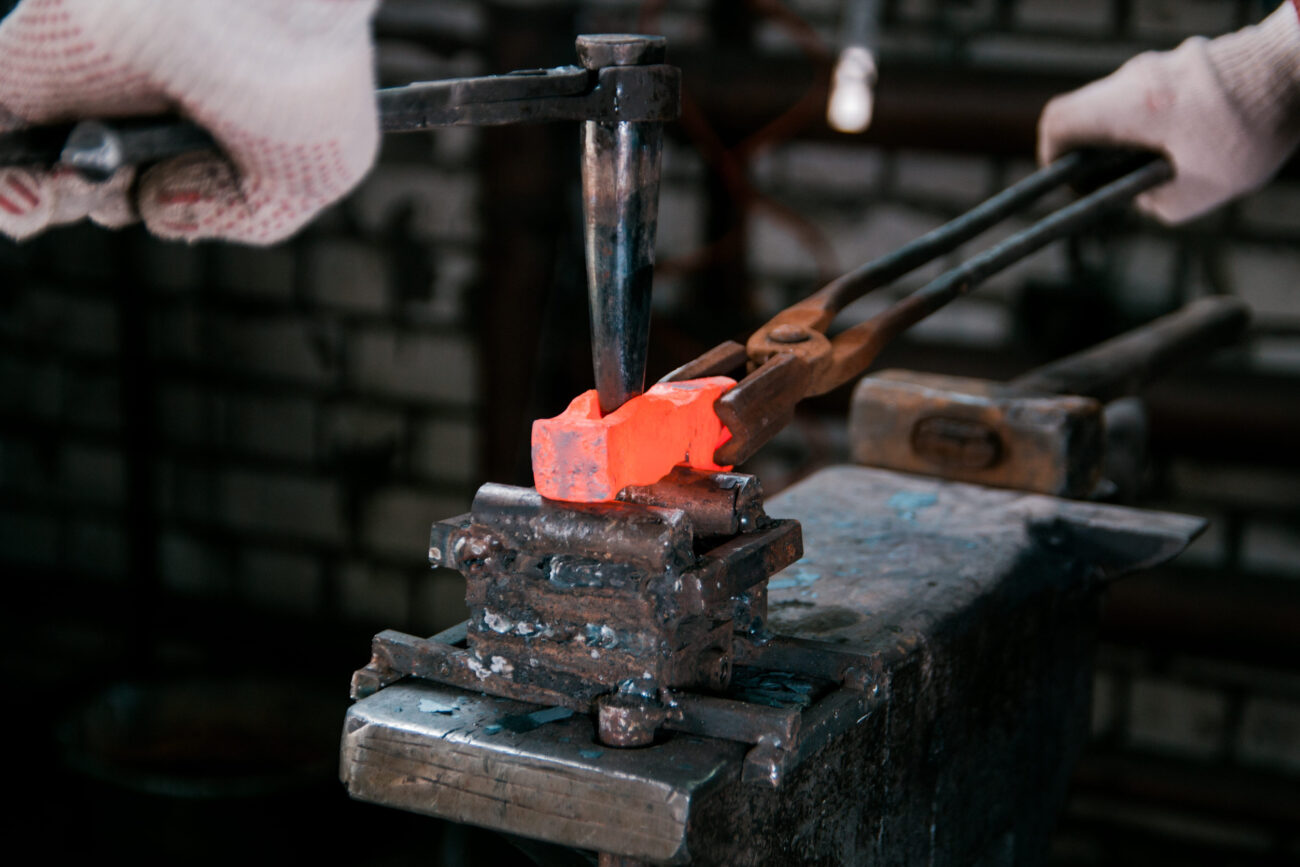 Hot orange metal being shaped in a blacksmith's hammering vise, with gloved hands and tongs in a workshop.
