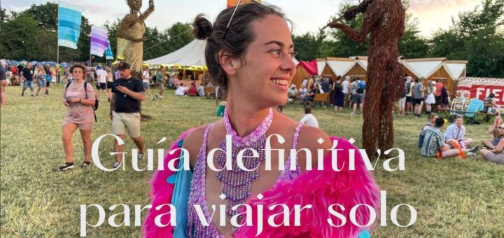 Smiling woman in a bright pink feathered festival outfit at an outdoor fair, with large white Spanish text over the image saying something about a solo travel guide.