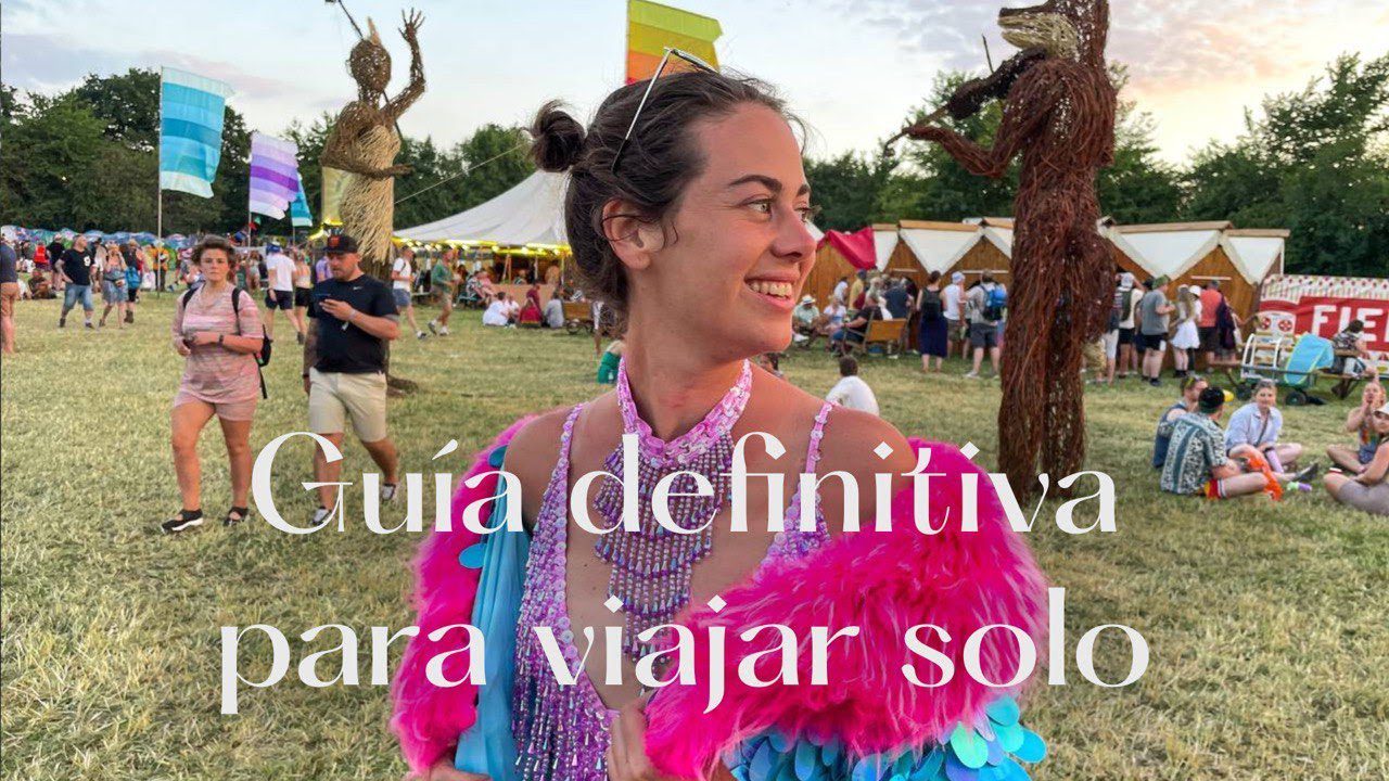 Guia-completa-sobre-viajes-en-solitario | Diario-ABC.Com Smiling woman in a bright pink feathered festival outfit at an outdoor fair, with large white Spanish text over the image saying something about a solo travel guide.