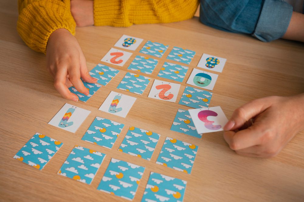 756595 - Warcks | Diario-ABC.Com Two people playing a card-matching game on a wooden table, blue cloud-pattern cards laid out in rows.