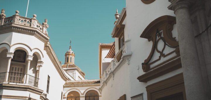 White Mediterranean-style buildings with arches and a domed tower against a turquoise sky in the background.