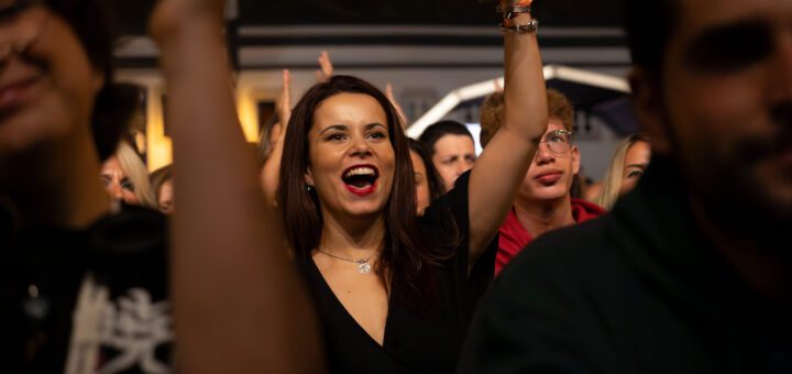 Young woman in a black top cheers with one arm raised, holding a blue cup, amid a cheering crowd at a night event.Good for a party or concert scene.