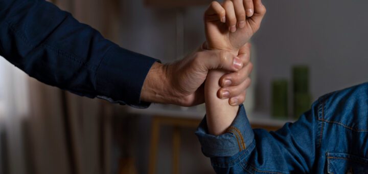 Adult hand firmly grips a child's forearm during an arm-wrestling-style grip in a home setting, suggesting guidance or restraint.