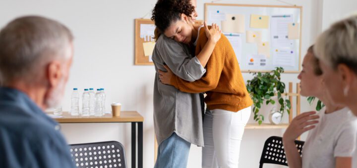 Two coworkers share a hug in a bright office as seated colleagues watch nearby.