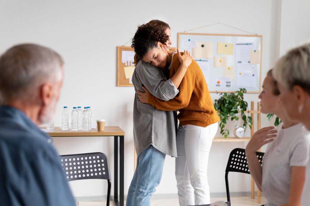 776376 - Mensalud | Diario-ABC.Com Two coworkers share a hug in a bright office as seated colleagues watch nearby.