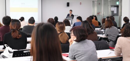 Speaker at a podium giving a presentation to a room full of attendees in a conference room with a projection screen.
