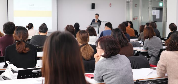 Speaker at a podium giving a presentation to a room full of attendees in a conference room with a projection screen.