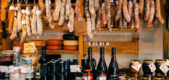 Rows of hanging cured sausages and salumi above a market stall, with bottles, jars, and cheeses on the wooden counter below