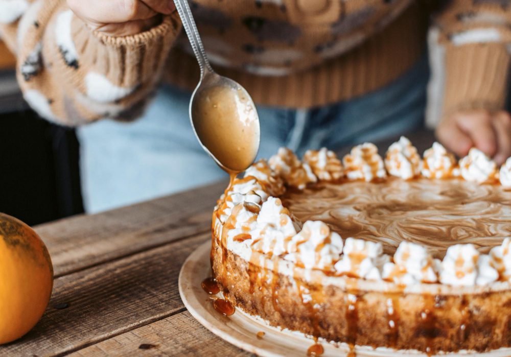 778458 - Longinos | Diario-ABC.Com Person in a cozy sweater drizzling caramel sauce over a circular cake with whipped-cream swirls on a wooden table.