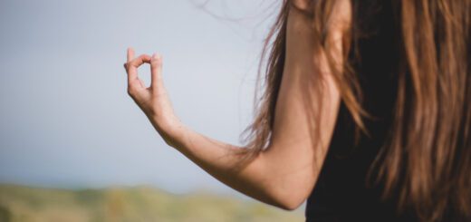 Back view of a person with long brown hair, outdoors in a field, making a circle with thumb and index finger (hand gesture)