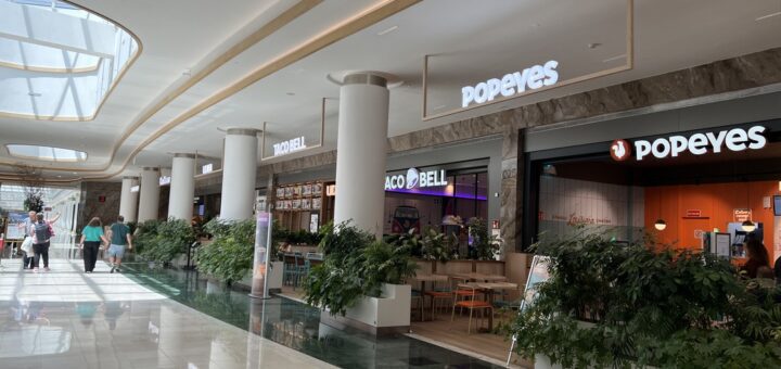 Bright mall corridor with white columns, potted plants, and fast-food storefronts including Popeyes and Taco Bell.
