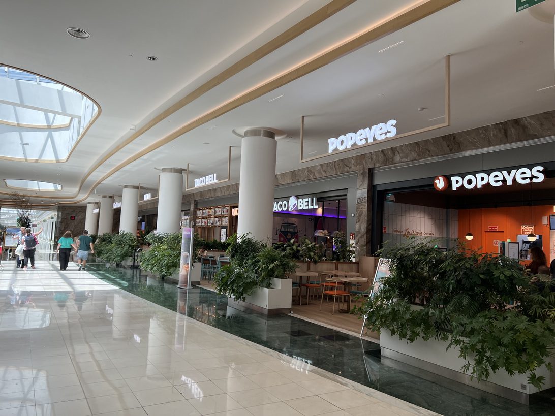 Gran Plaza 2 Restauración - IP | Diario-ABC.Com Bright mall corridor with white columns, potted plants, and fast-food storefronts including Popeyes and Taco Bell.