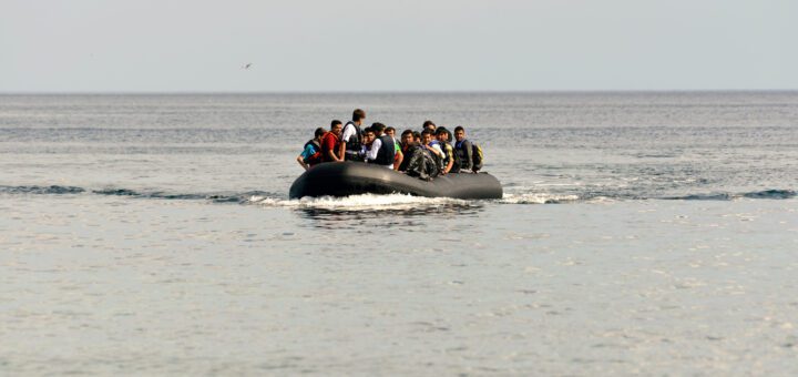 Crowded inflatable boat with passengers wearing life jackets on a calm sea near the horizon.