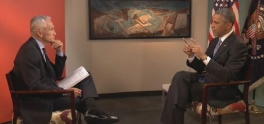 Two men in suits sit facing each other in a studio interview setting, one holding papers with American flags and a painting in the background.