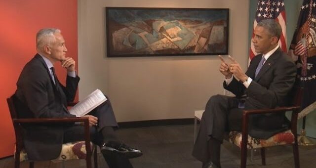 Two men in suits sit facing each other in a studio interview setting, one holding papers with American flags and a painting in the background.