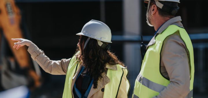 Two construction workers in hard hats and high-vis vests discuss plans at a building site; one points while the other holds blueprints.