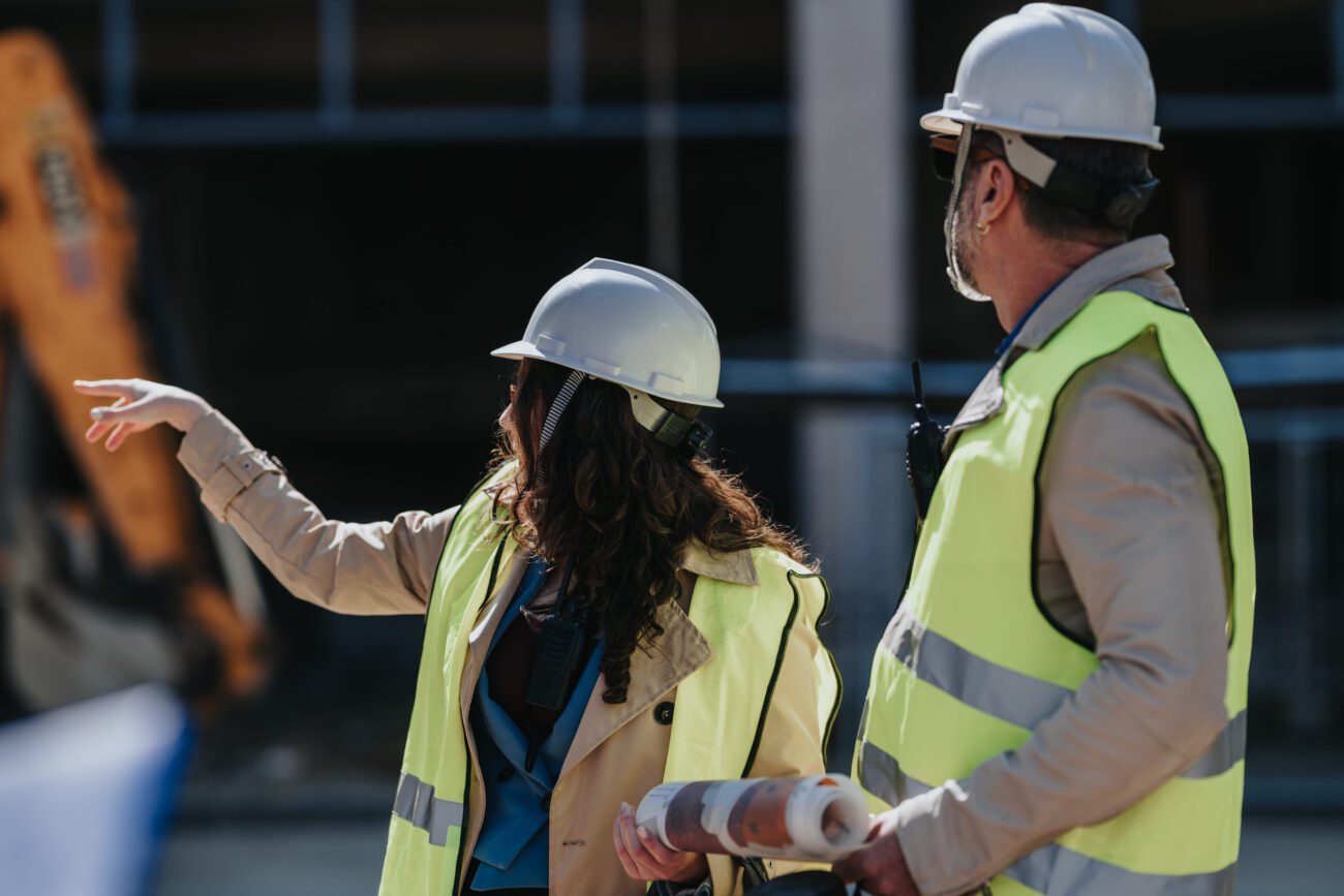 Two construction workers in hard hats and high-vis vests discuss plans at a building site; one points while the other holds blueprints.
