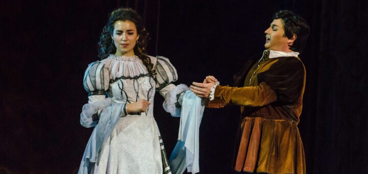 Stage performance: a woman in a white, ornate period dress and a man in a brown velvet tunic on stage, holding hands