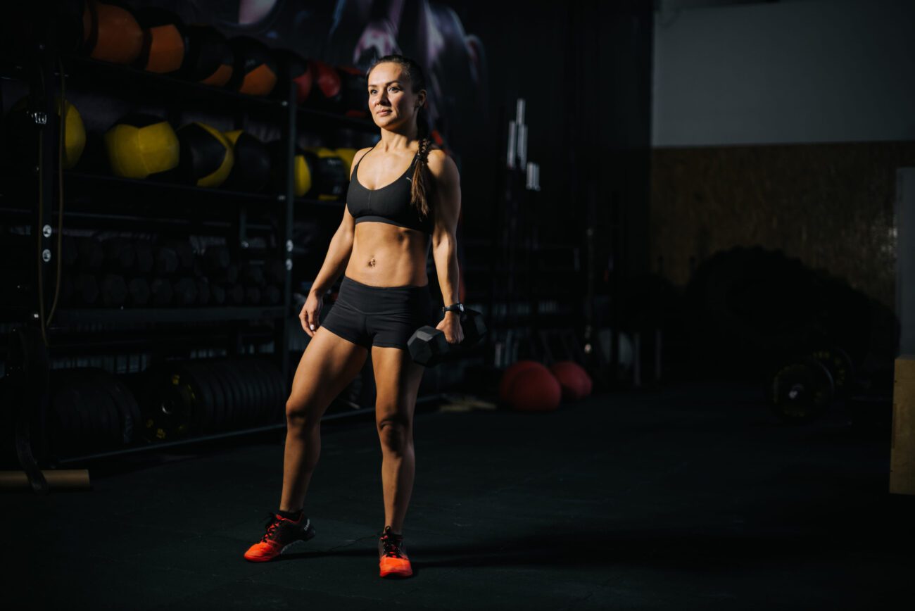 780816 isobiv fold | Diario-ABC.Com Woman in a gym standing with a dumbbell in her right hand, wearing a black sports bra, black shorts, and bright orange sneakers, lit from the side.