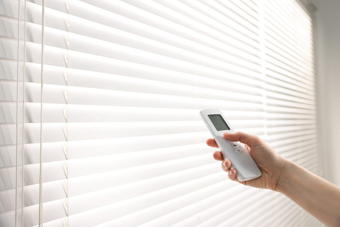 781918 Cortimarket | Diario-ABC.Com Hand holding a white remote control aimed at white horizontal window blinds