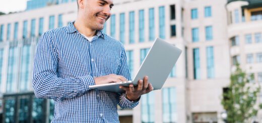 Man in a blue checkered shirt smiles while using a silver laptop outdoors in an urban area with modern glass buildings behind him.