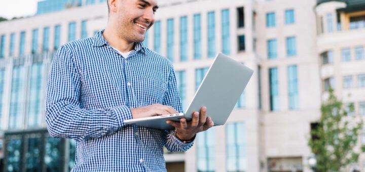 Man in a blue checkered shirt smiles while using a silver laptop outdoors in an urban area with modern glass buildings behind him.
