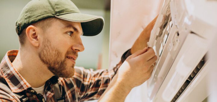 Male technician in a green cap repairing a large wall-mounted control panel on a machine/breakroom appliance.
