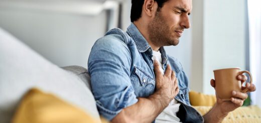Man on a couch clutching his chest with a pained expression while holding a mug in one hand.