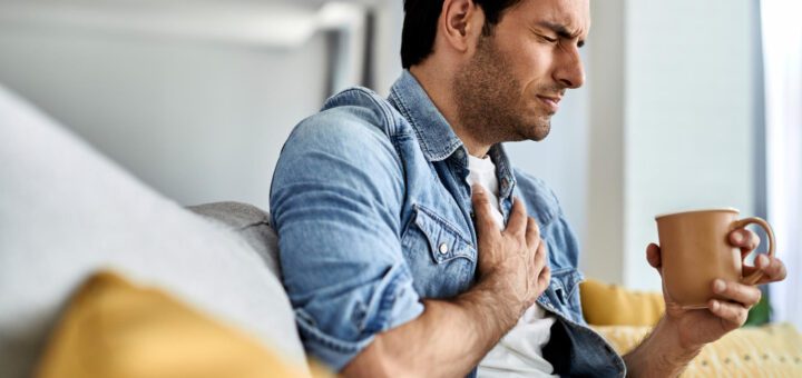 Man on a couch clutching his chest with a pained expression while holding a mug in one hand.