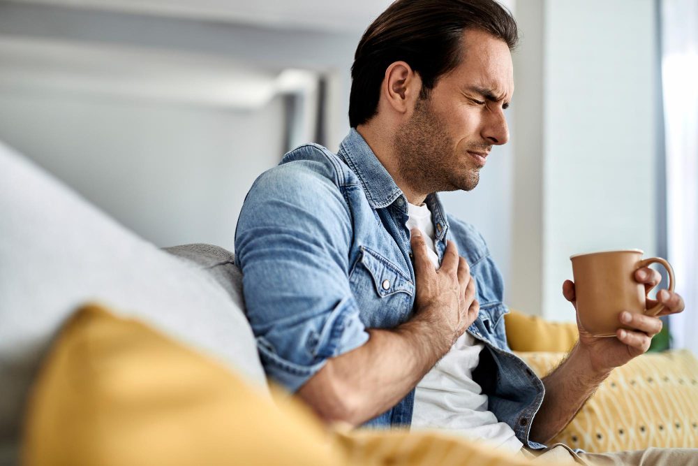 782545 - Clínica Cardiovascular Dr Valderrama | Diario-ABC.Com Man on a couch clutching his chest with a pained expression while holding a mug in one hand.
