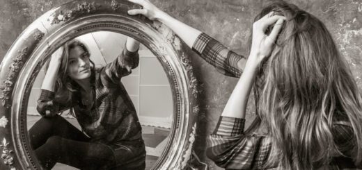 Woman in a plaid shirt sits on the floor and studies her reflection in a large ornate round mirror.