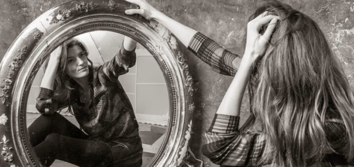 Woman in a plaid shirt sits on the floor and studies her reflection in a large ornate round mirror.