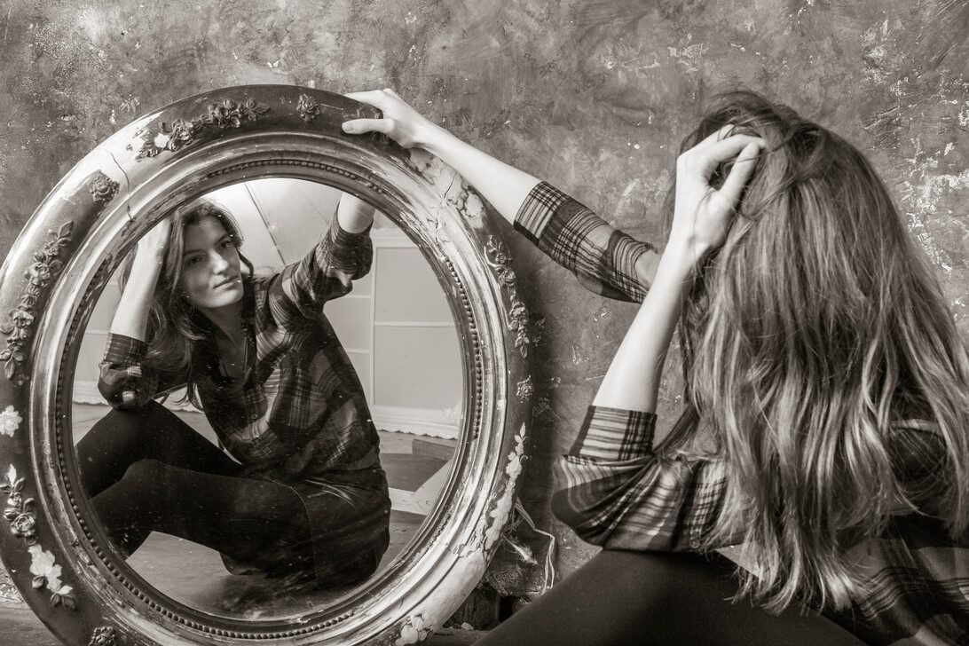 Young longhaired beautiful woman sitting in front of the vintage mirror | Diario-ABC.Com Woman in a plaid shirt sits on the floor and studies her reflection in a large ornate round mirror.