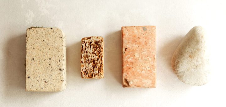 Four soap bars in a row: beige speckled loaf, small brown textured bar, pink rectangular soap, and a pale pebble-shaped bar on a light background.