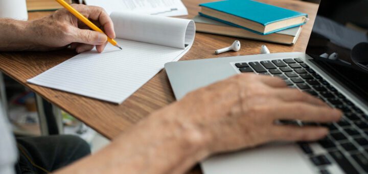 Close-up of hands writing with a pencil in a lined notebook on a wooden desk, with a laptop, earbuds, and stacked books nearby.
