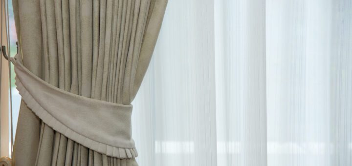 Beige heavy drapes tied back with a matching tieback beside sheer white curtains behind.