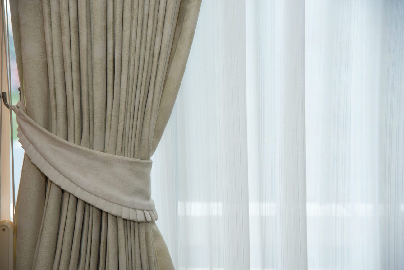 Beige heavy drapes tied back with a matching tieback beside sheer white curtains behind.