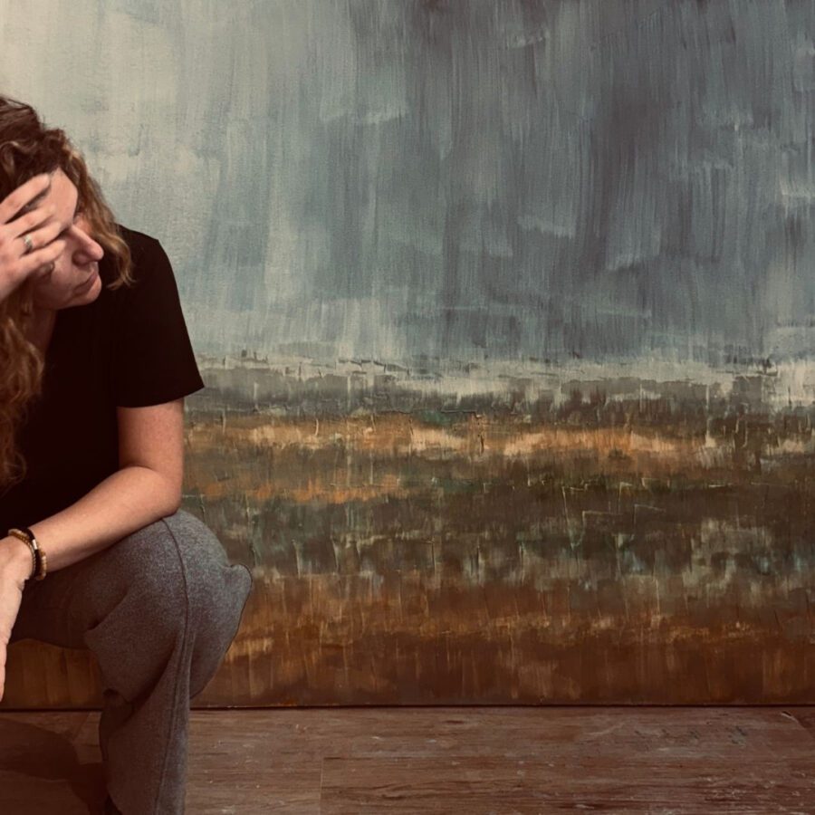 Berta Giner - IP | Diario-ABC.Com Woman with long curly hair sits with her head resting in her hand, looking down, against a textured blue-gray wall.