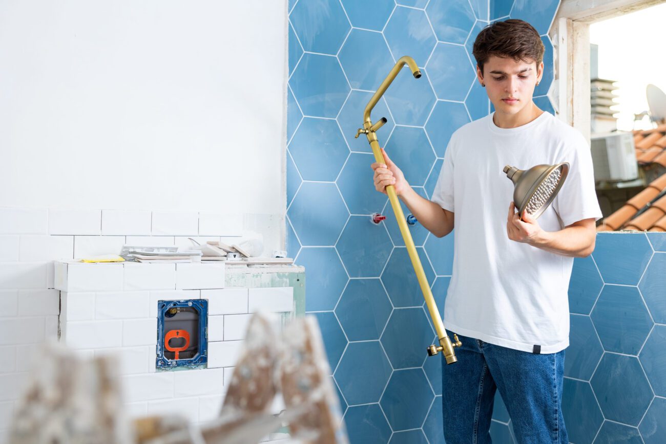 Focused guy assembling shower set before fixing to wall | Diario-ABC.Com Young man installing a brass shower pipe and spray head in a bathroom under renovation.