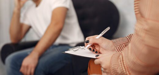 Close-up of a person writing on a clipboard with a pen, while a man sits in the background seated in a chair.