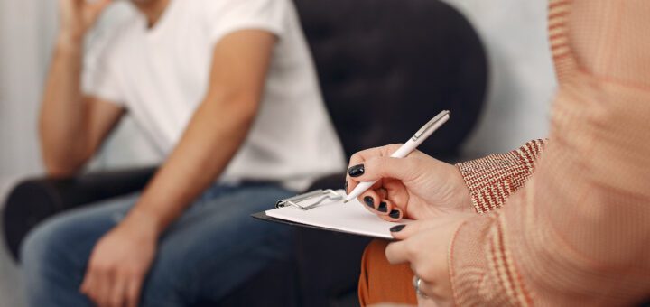 Close-up of a person writing on a clipboard with a pen, while a man sits in the background seated in a chair.