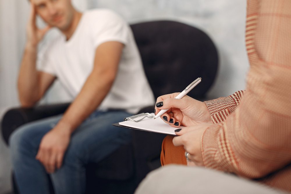 Close-up of a person writing on a clipboard with a pen, while a man sits in the background seated in a chair.