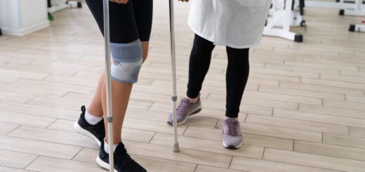 Person using forearm crutches and wearing a knee brace during a physical therapy session, guided by a clinician in a gym.