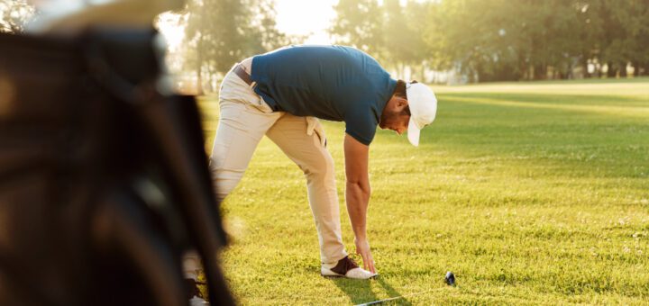 Man in a blue shirt and beige pants lines up a golf tee, placing a ball on the ground beside a golf bag in a sunny park course.