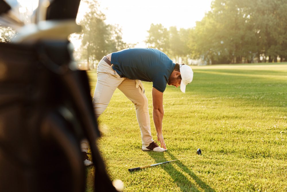 784291 - Black Iron | Diario-ABC.Com Man in a blue shirt and beige pants lines up a golf tee, placing a ball on the ground beside a golf bag in a sunny park course.