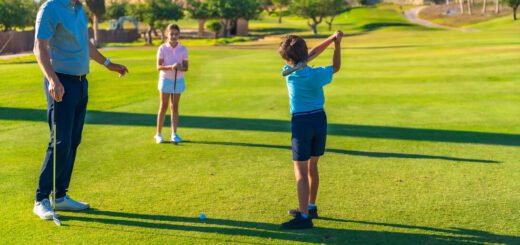 Boy in blue shirt swings a golf club while a man watches on a sunny course; a girl stands with a club in the background.
