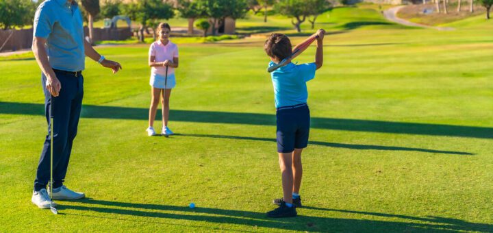 Boy in blue shirt swings a golf club while a man watches on a sunny course; a girl stands with a club in the background.